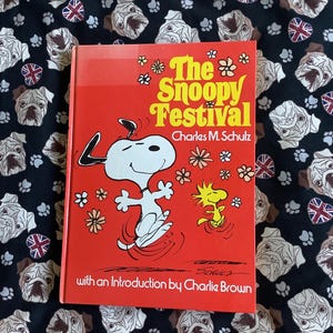 May include: A red book titled "The Snoopy Festival" by Charles M. Schulz, featuring Snoopy and Woodstock. The cover has a cartoon illustration of Snoopy dancing. The background is a black fabric with bulldog and Union Jack patterns.