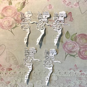 Könnte beinhalten: Fünf weiße Papier-Ausschnitt-Feenfiguren mit filigranen Flügel- und Kleid-Details, jede in einer Pose. Die Feen sind vor einem floralen und spitzenartigen Hintergrund mit Textfragmenten platziert. Der Text enthält die Wörter "SONG DA FODIL" und "ONG O LOVE FA".