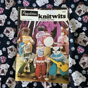 May include: A vintage knitting pattern book titled "Knitwits" featuring a band of knitted animal characters. The cover shows a group of knitted animals playing musical instruments. The background is a dark fabric with a pattern of white pugs and Union Jack flags.