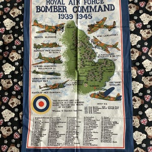 Pode incluir: Um pano de cozinha azul marinho com um mapa do Reino Unido com ilustrações de aviões do Royal Air Force Bomber Command de 1939-1945. O pano inclui texto e uma lista de aeródromos.