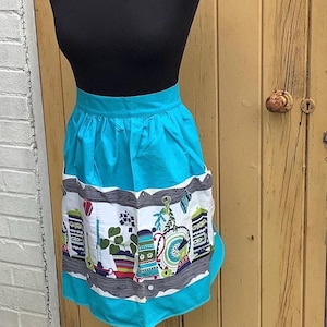 May include: A turquoise apron with a black top, displayed on a mannequin. The apron features a white panel with colorful illustrations of teapots, plants, and other kitchen items. The apron is approximately 61 cm long.