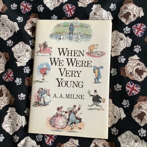 Op de afbeelding: Een boek met de titel "When We Were Very Young" van A.A. Milne, met kleurrijke illustraties op de omslag. Het boek ligt op een zwarte stof met bulldoggezichten, pootafdrukken en Union Jack-vlaggen.