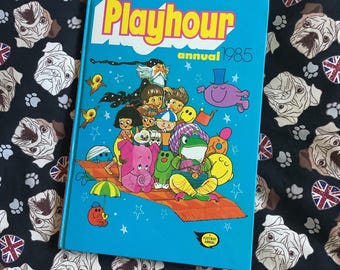 May include: A blue book with the title "Playhour Annual 1985" and a colourful illustration of cartoon characters on the cover.