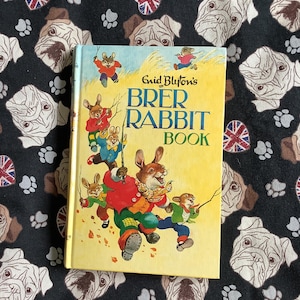 May include: A vintage children's book titled "Brer Rabbit Book" by Enid Blyton. The cover features illustrations of rabbits in colorful clothing. The book rests on a black fabric patterned with bulldog faces, paw prints, and Union Jack flags.