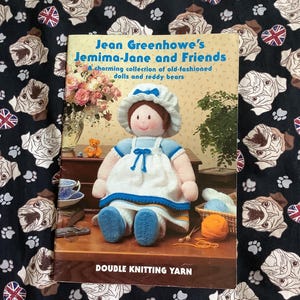 May include: A book titled "Jean Greenhowe's Jemima-Jane and Friends" featuring a knitted doll and teddy bear. The book cover displays a doll in a blue and white dress. The background is a black fabric with pug dog and Union Jack patterns.