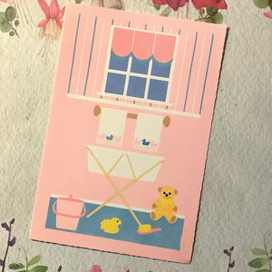 May include: A pink greeting card with a baby-themed illustration. The card features a window, towels, a baby tub, a teddy bear, a duck, and a pink bucket. The background has a floral design.