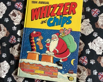 May include: A yellow and red comic book titled "Whizzer and Chips 1984 Annual". The cover features a cartoon illustration of Santa Claus climbing down a chimney with a sack of presents. A snake is also on the chimney.