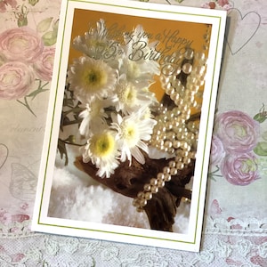 May include: A greeting card with a white border and green trim features white flowers, pearls, and the text "Wishing you a Happy Birthday." The card's background is a warm orange color.