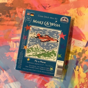 May include: Cross stitch mini kit featuring a red airplane flying over green mountains. The kit includes DMC stranded cotton, a needle, and instructions. The kit is 7.5 cm by 7.5 cm and supports Make-A-Wish.