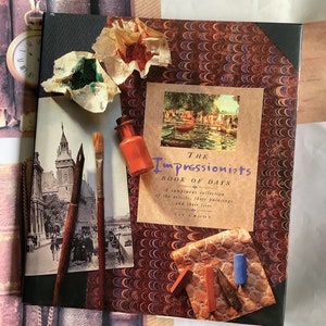 May include: A book titled "The Impressionists Book of Days" with a marbled cover. The book features a painting of a canal scene with boats and trees. The cover also includes a paintbrush, a small bottle of paint, and a few pieces of chalk.