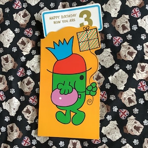 Puede incluir: Una tarjeta de cumpleaños con un personaje de dibujos animados sosteniendo un globo rosa y un regalo. La tarjeta es naranja y dice "HAPPY BIRTHDAY NOW YOU ARE 3". El fondo es una tela negra con caras de pug.