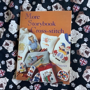 May include: An orange book titled "More Storybook Cross-stitch" featuring cross-stitch designs. It rests on black fabric adorned with white bulldog faces, paw prints, and Union Jack flags. The book showcases various cross-stitch projects.
