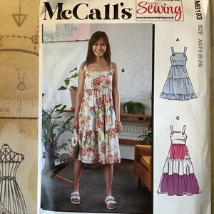 May include: McCall's sewing pattern M8193 for sizes A5/F5 (6-24) features three dress styles. The front of the pattern shows a woman wearing a floral print dress with a tiered skirt. The back of the pattern shows two additional dress styles: a blue dress with a tiered skirt and a pink and purple dress with a tiered skirt.