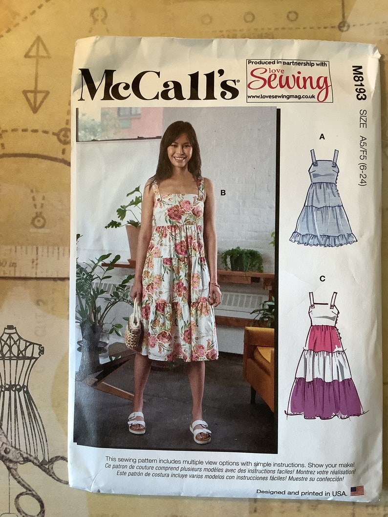 May include: McCall's sewing pattern M8193 for sizes A5/F5 (6-24) features three dress styles. The front of the pattern shows a woman wearing a floral print dress with a tiered skirt. The back of the pattern shows two additional dress styles: a blue dress with a tiered skirt and a pink and purple dress with a tiered skirt.