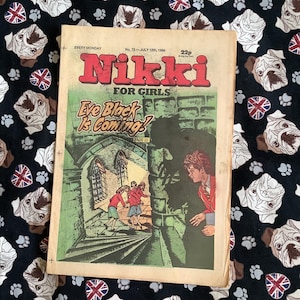 May include: A well-worn copy of the comic "Nikki FOR GIRLS" from July 12th, 1986, featuring the story "Eve Black Is Coming!". The cover illustration shows figures in a gothic setting. The comic rests on a black fabric patterned with bulldog faces and Union Jack flags.