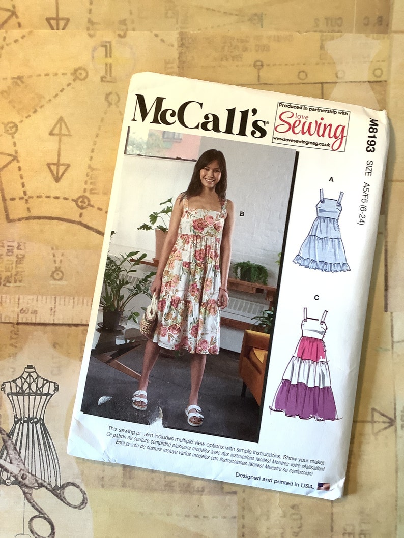 May include: McCall's Sewing pattern M8193, size A5/F5 (6-24), featuring three different styles of dresses. The first dress is a floral print, the second is a light blue tiered dress, and the third is a pink, white, and purple tiered dress. The pattern includes simple instructions and multiple view options.