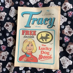 May include: A vintage "Tracy" magazine for girls, dated November 6, 1982, with a cover featuring a blonde girl and the text "The Lucky Dog Brooch." The magazine is set against a black fabric patterned with bulldog faces and Union Jack flags.