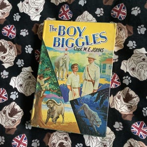 May include: A vintage paperback book titled "The Boy Biggles" by Capt. W.E. Johns. The cover features illustrations of a boy, an elephant, a buffalo, and a tiger.