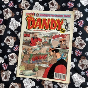 May include: A vintage comic book cover featuring a red and yellow illustration of "The Dandy" with the text "Free Desperate Dan Button Badge" and "Fun for boys and girls!" The comic book cover is dated November 1996 and costs 42p. The comic book cover features a cartoon illustration of Desperate Dan, a strong man, with the text "Desperate Dan" and "Creak!"