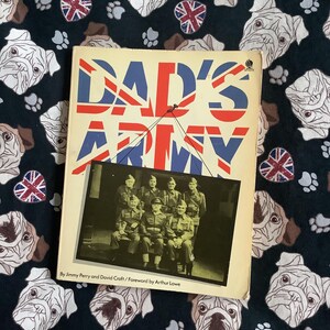 May include: A vintage book titled "Dad's Army" with a Union Jack design on the cover. The cover features a black and white photo of a group of men in military uniforms. The book is set against a dark background with bulldog and Union Jack patterns.