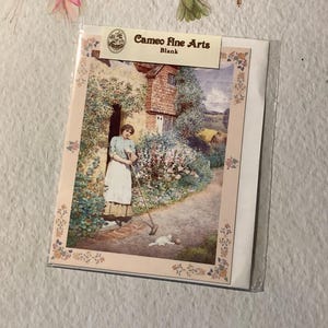 May include: A blank greeting card featuring a vintage painting of a woman in a garden with a white cat. The card is in a clear plastic sleeve. The card has the text "Cameo Fine Arts" and "Blank".