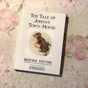 Op de afbeelding: Een hardcover boek getiteld "The Tale of Johnny Town-Mouse" van Beatrix Potter. De omslag toont een illustratie van een muis met een koffer. Het boek is een nieuwe kleurenreproductie van F. Warne & Co.