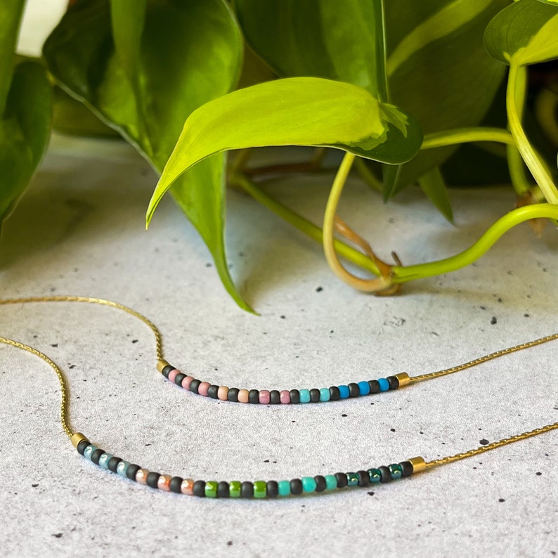 May include: Two delicate gold necklaces with colorful beads. The top necklace has pink, blue, and black beads. The bottom necklace has green, blue, and black beads.