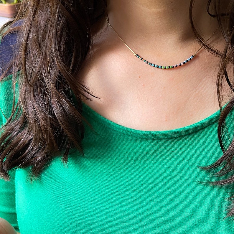 May include: A woman wearing a green shirt with a delicate gold chain necklace featuring colorful beads.