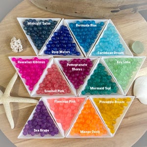 May include: Assortment of colorful translucent beads in triangular dishes. Each dish is labeled with a name, such as "Midnight Swim", "Bermuda Blue", "Deep Waters", "Hawaiian Hibiscus", "Pomegranate Shores", "Caribbean Dream", "Key Lime", "Seashell Pink", "Flamingo Pink", "Mermaid Teal", "Pineapple Breeze", "Sea Grape", and "Mango Oasis".