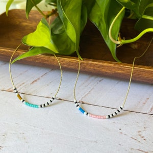 May include: Two gold necklaces with colorful beaded accents. The necklaces feature black and white beads with a pop of color in the center. One necklace has a turquoise accent and the other has a pink accent.