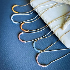 May include: A collection of delicate necklaces with a crescent-shaped design. Each necklace features a row of small, colorful beads in shades of orange, white, peach, blue, and pink. The beads are strung on thin, gold and silver-colored cords.
