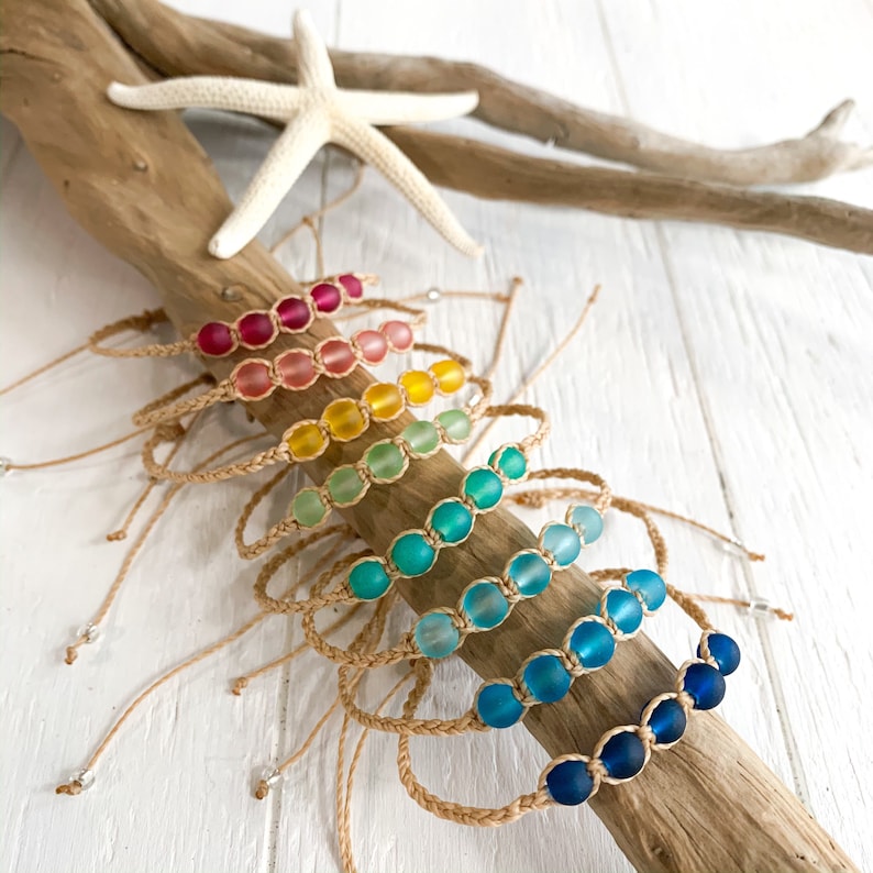 May include: A set of eight colorful bracelets made with small glass beads and natural twine. The bracelets are arranged in a rainbow pattern, with each bracelet featuring a different color of beads.