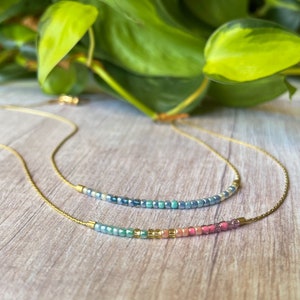 May include: Two delicate gold necklaces with iridescent beads. The top necklace features blue and green beads, while the bottom necklace has a rainbow of colors, including pink, blue, and gold.