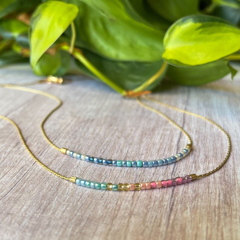 May include: Two delicate gold necklaces with iridescent beads. The top necklace features blue and green beads, while the bottom necklace has a rainbow of colors, including pink, blue, and gold.