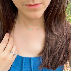 May include: A gold chain necklace with a rainbow of tiny beads. The necklace is worn by a person with long brown hair and a blue shirt.