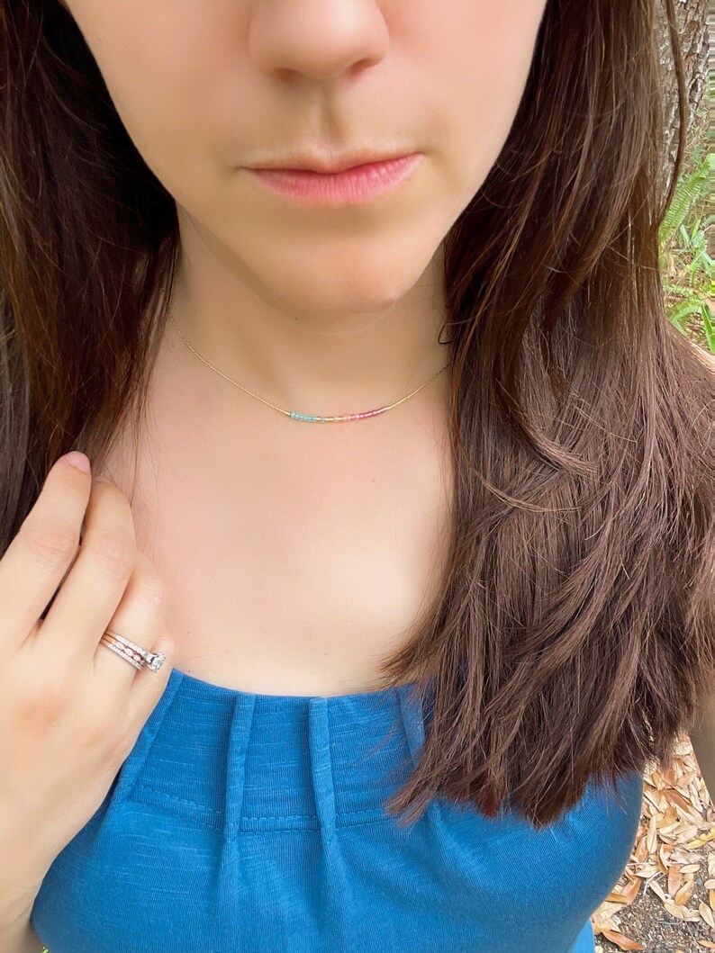 May include: A gold chain necklace with a rainbow of tiny beads. The necklace is worn by a person with long brown hair and a blue shirt.