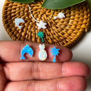 May include: A set of three opal earrings featuring a dolphin, pineapple, and palm tree design. The earrings are a light blue, white, and iridescent color.