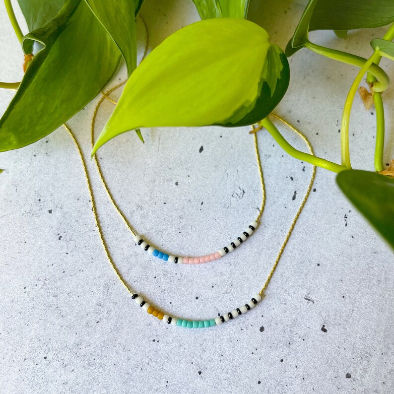 May include: Two delicate gold necklaces with colorful beaded charms. The top necklace features black and white beads with pink, blue, and white beads in the center. The bottom necklace features black and white beads with yellow, blue, and white beads in the center.