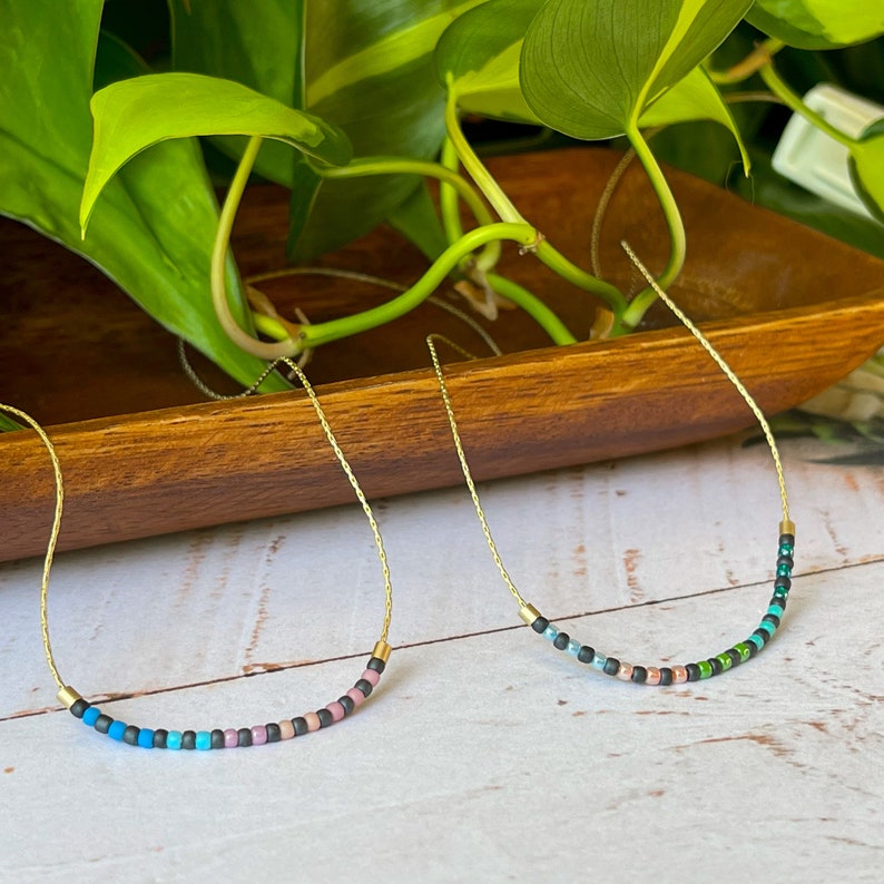 May include: Two delicate gold necklaces with colorful beaded accents. The necklaces are arranged on a wooden surface with green foliage in the background.