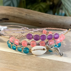 May include: Three layered bracelets with pink, purple, and blue beads. The bracelets are made with a beige cord and have a seashell charm and a silver seahorse charm.