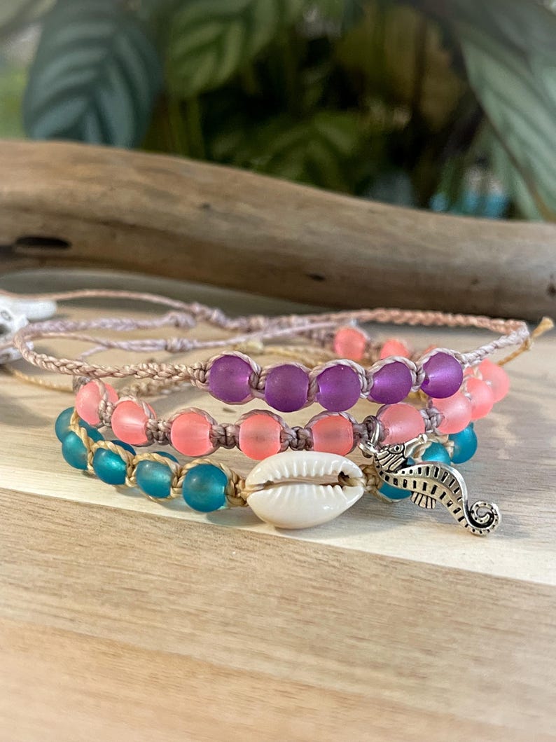 May include: Three layered bracelets with pink, purple, and blue beads. The bracelets are made with a beige cord and have a seashell charm and a silver seahorse charm.