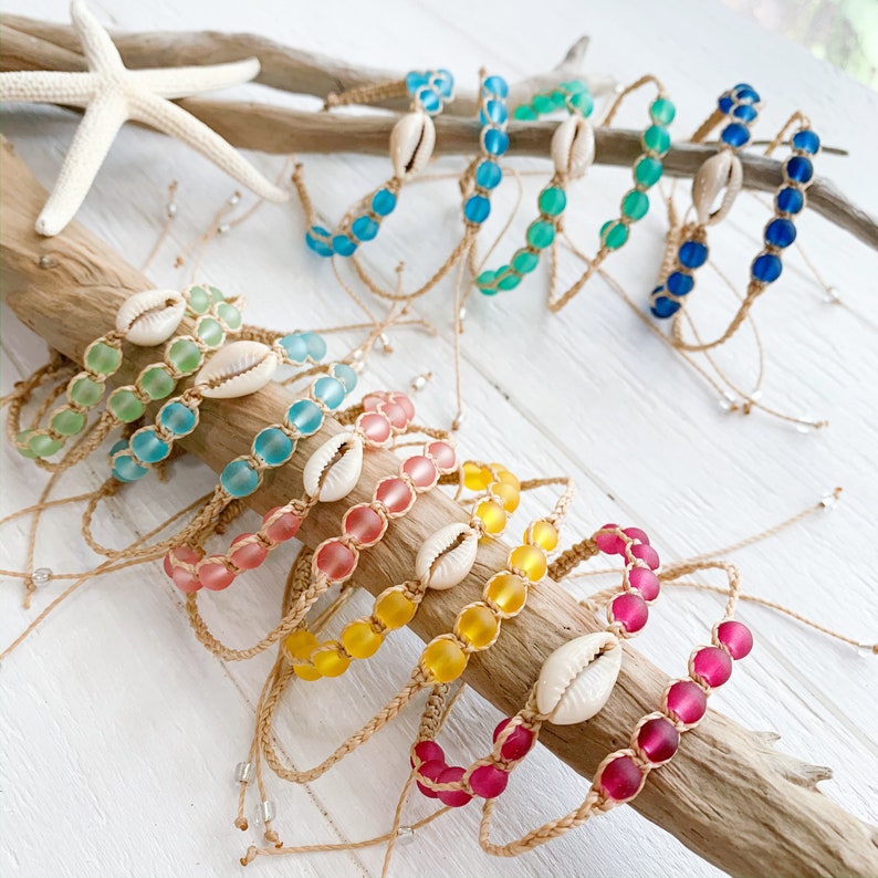 May include: A set of colorful bracelets made with glass beads and shell charms. The bracelets are strung on a natural cord and come in a variety of colors, including blue, green, pink, yellow, and red.