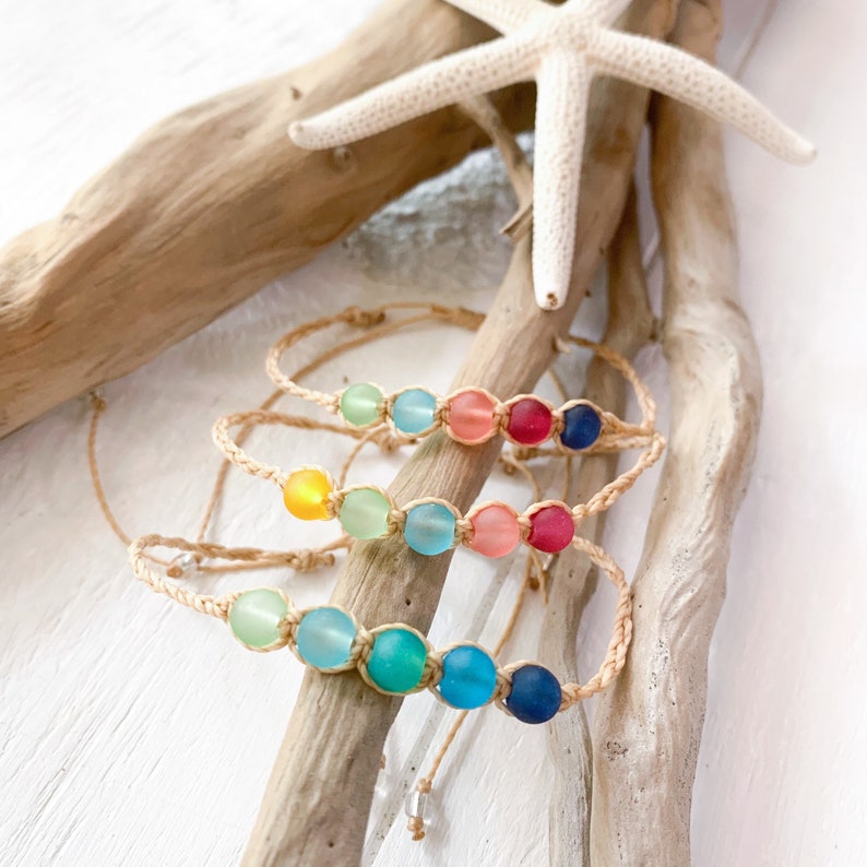 May include: Four bracelets with different colored glass beads on a natural beige background. The beads are in shades of blue, green, yellow, pink, and red. The bracelets are made with a natural fiber cord.