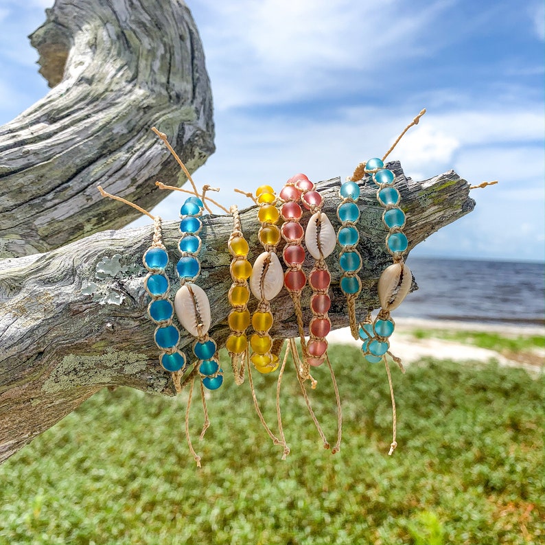 May include: A set of six colorful beaded bracelets with shell accents. The bracelets are made with blue, yellow, and pink beads and are strung on natural twine. The bracelets are displayed on a weathered piece of driftwood.
