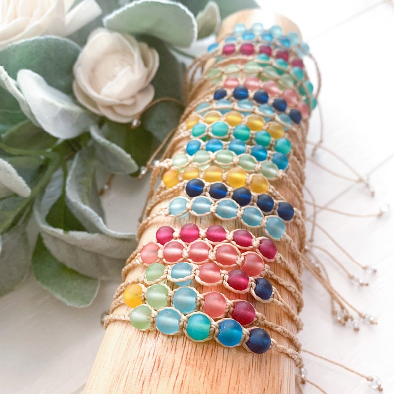 May include: A stack of colorful beaded bracelets with a variety of colors including blue, green, yellow, pink, and red. The bracelets are made with small glass beads and natural string.
