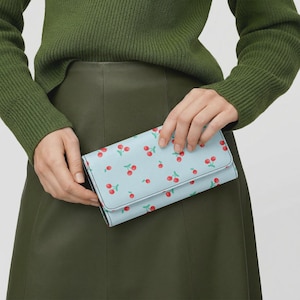 May include: A rectangular wallet in light blue featuring a repeating pattern of small red cherries with green stems. The wallet has a fold-over flap closure and is held in place, showcasing the exterior design and the smooth texture of the material.