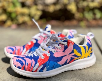 Puede incluir: Un par de zapatillas coloridas con un estampado floral. Los zapatos tienen suela y cordones blancos, con un diseño de hojas rosas, rojas, amarillas y azules sobre un fondo azul. Las zapatillas están sobre una superficie de hormigón.