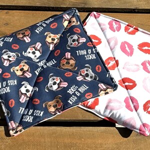 May include: Two pot holders, one with a blue background and a pattern of white dogs with pink tongues and the text "100% Kiss a Bull", and the other with a white background and a pattern of pink lips.