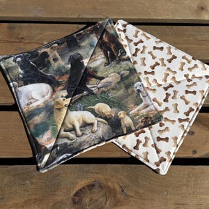 May include: Two square quilted pot holders. One features a print of various dog breeds in a natural setting. The other has a white background with a pattern of brown dog bones. The pot holders are on a wooden surface.