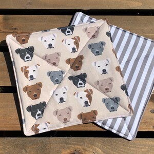 May include: Two square fabric pot holders. One has a repeating pattern of dog faces in various colours on a beige background. The other has a grey and white striped pattern. Both have white trim.
